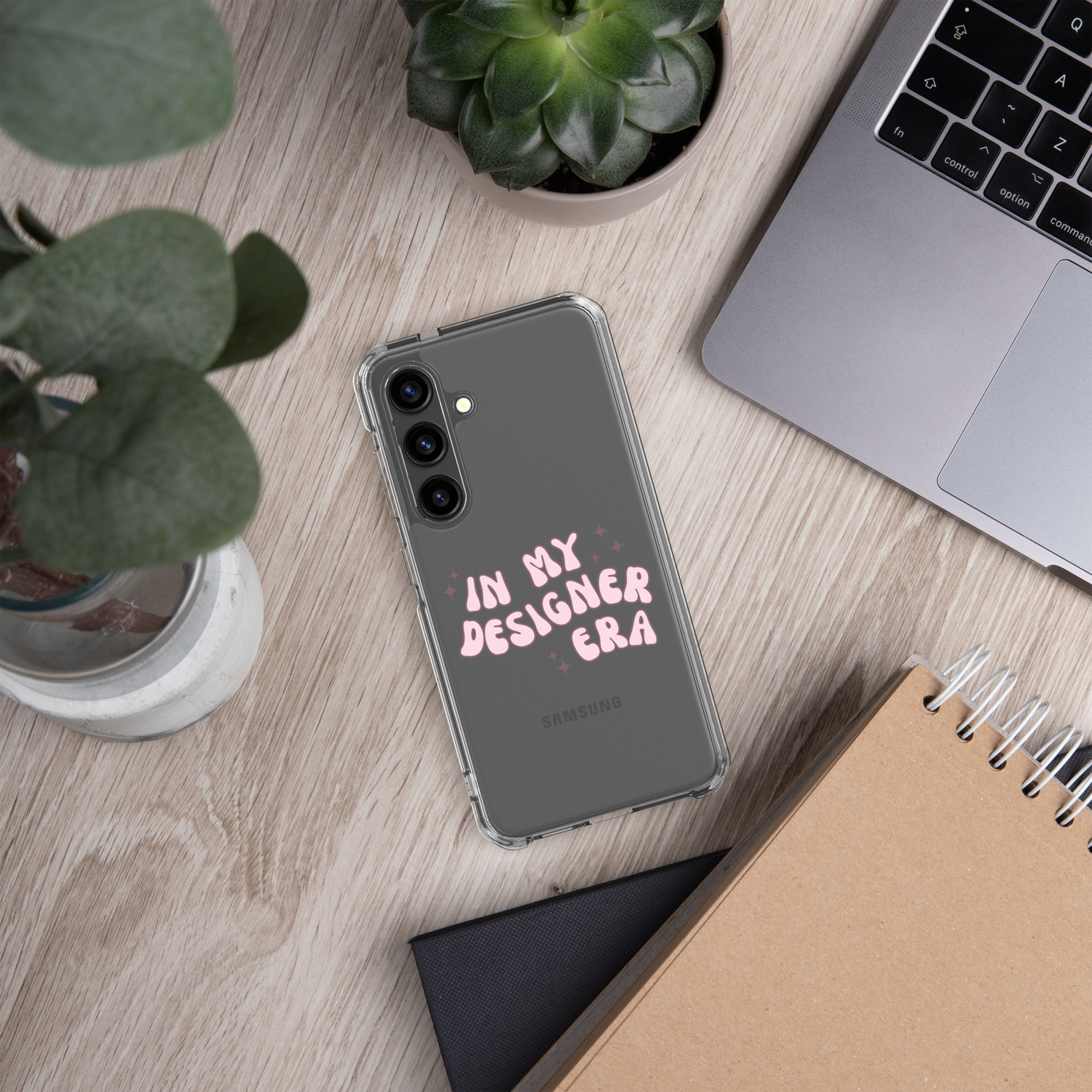 Wood desk with plants, laptop, notebooks, and phone artfully displayed. Phone has a clear case that says In My Designer Era in puffy pink letters